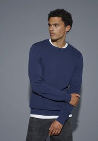 Navy blue textured sweater with a crew neck and ribbed cuffs, layered over a white shirt, paired with dark jeans.