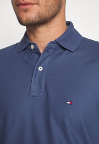 Polo bleu marine en tissu texturé, avec un col à deux boutons et un petit logo rouge, blanc et bleu sur la poitrine gauche.
