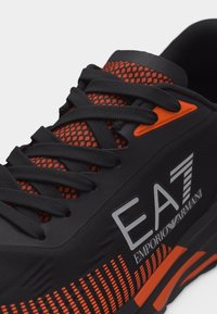 Μαύρο και πορτοκαλί αθλητικό παπούτσι με μαύρα κορδόνια, δίχτυ στη γλώσσα, λογότυπο EA7 Emporio Armani και διακοσμητική πορτοκαλί σόλα με σχέδιο.