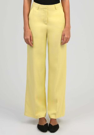 Femme portant un pantalon jaune à jambes larges, un haut noir et des chaussures noires pointues, debout devant un fond clair uni.