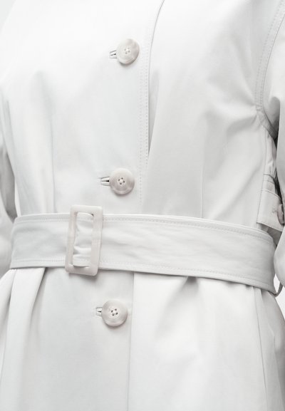 Fehér ballonkabát övvel, nagy gombokkal és sima textúrával. Az anyag könnyűnek és vízlepergetőnek tűnik.