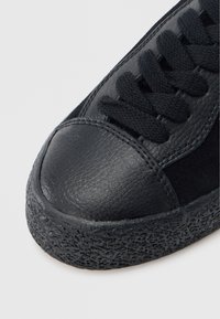 Czarne trampki z teksturowaną gumową podeszwą, gładkim skórzanym noskiem i sznurowadłami z tkaniny. Projekt ma niski krój i płaski profil.