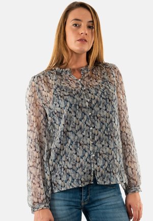 Blusa trasparente con un motivo astratto blu e beige, colletto arricciato, maniche lunghe e chiusura frontale con bottoni. Abbinata a jeans blu.