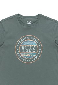 Camiseta de algodón gris con cuello redondo que presenta un logo circular en olas naranjas y azules. El texto incluye "Calidad Premium" y "Billa Bong."