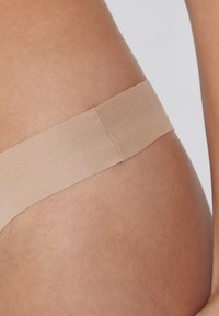 Beiger Tanga aus weichem Stoff mit glatter Textur. Der Bund ist elastisch und nahtlos, was einen diskreten Sitz auf der Haut gewährleistet.