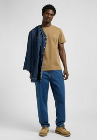 Beige Baumwoll-T-Shirt mit dezentem Logo, kombiniert mit blauem Jeansjacke und lässigen blauen Jeans. Braune Schnürstiefel runden den Look ab.