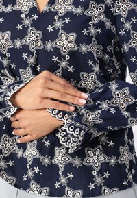 Blusa blu navy con ricami floreali bianchi. Presenta dettagli intricati sulle maniche e una chiusura con bottoni, realizzata in tessuto leggero.