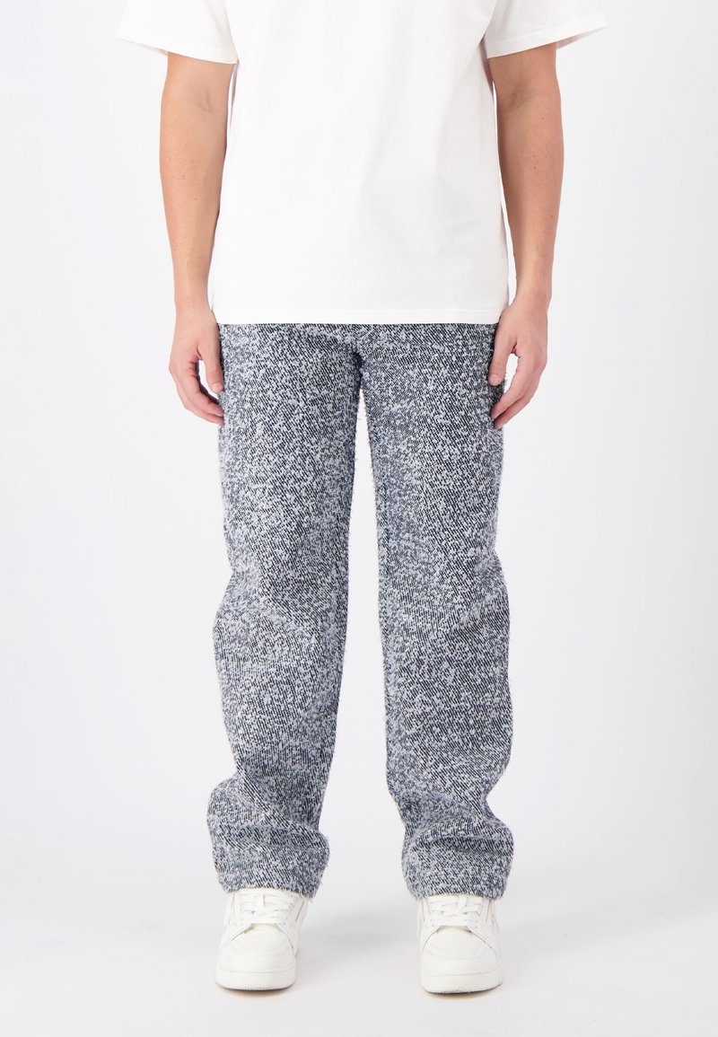 Grijs en wit gespikkelde sweatpants van een textuurgebreid materiaal. Losse pasvorm met een taps toelopende boord bij de enkels, gedragen met witte sneakers.