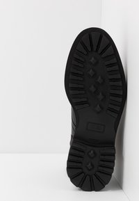 Semelle de botte en caoutchouc noir avec un motif de crampons profonds et une surface texturée, présentant des grips en forme de diamant et un bord lisse et arrondi.