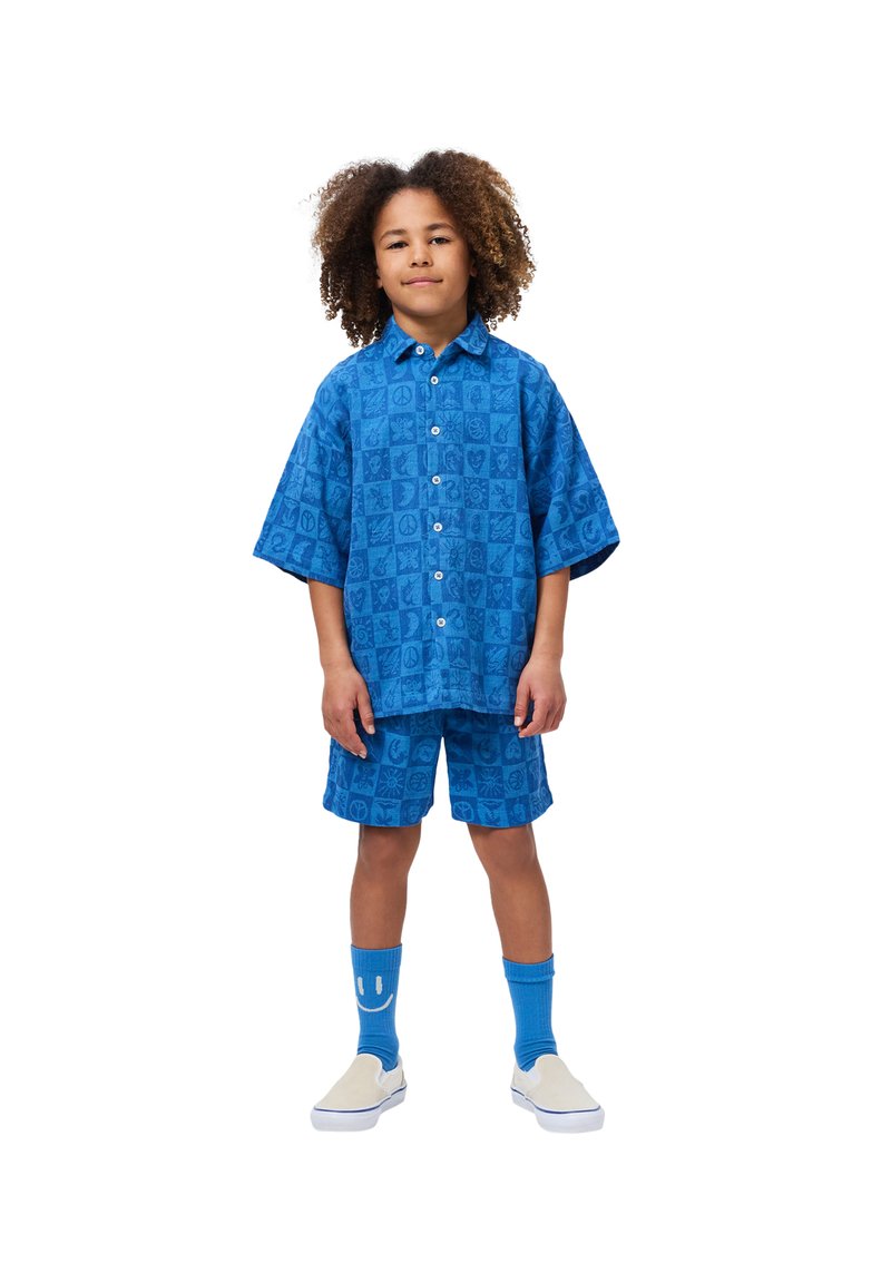 Σετ μπλε κοντομάνικης μπλούζας και σορτς με σχέδιο τετραγώνων που διαθέτουν σύμβολα. Συμπληρώνεται από μπλε κάλτσες και παπούτσια χωρίς κορδόνια.