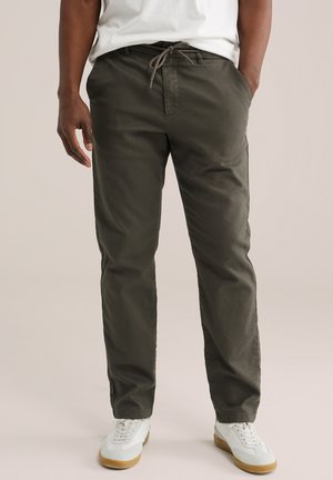 Uomo che indossa pantaloni verde oliva con coulisse e sneakers bianche, in piedi con una mano in tasca davanti a uno sfondo semplice.