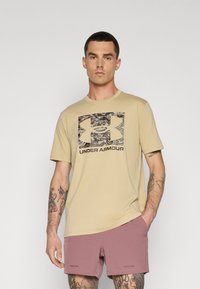 Camiseta beige con un gráfico de camuflaje y el logo de Under Armour. Combinada con pantalones cortos deportivos rosas, mostrando tatuajes visibles en los brazos.