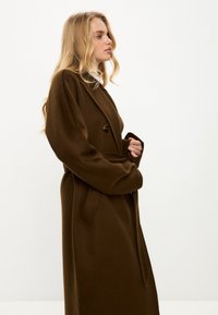 Brauner Wollmantel mit strukturiertem Finish, ausgestattet mit einem einzigen Knopf, tiefen Taschen und einer lockeren Silhouette. Lange Ärmel und ein Wickeldesign.