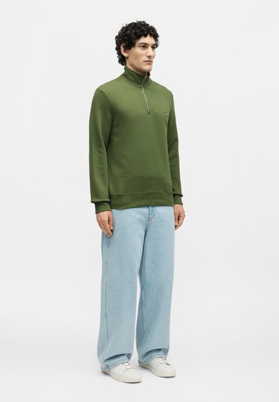 Άνδρας που φοράει μια ελιά-πράσινη μπλούζα με φερμουάρ μέχρι το στήθος, ανοιχτό μπλε τζιν με φαρδιά πόδια και λευκά αθλητικά παπούτσια, στέκεται μπροστά σε απλό φόντο.