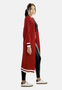 Larga chaqueta de punto roja con un corte holgado, que presenta puños a rayas blancas y rojas, combinada con leggings negros y zapatillas blancas.