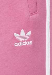 Die rosa Jogginghosen aus Stoff von Adidas verfügen über ein weiß gesticktes Logo mit drei blattähnlichen Designs und zwei kontrastierenden weißen Seitenstreifen.