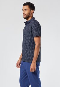 Lyhythihainen paita tummansinisestä kankaasta, jossa on geometrinen valkoinen kuvio, kaulus ja rento mitoitus, yhdistettynä sinisiin housuihin.