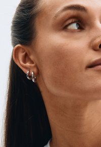 Hõbedased metallist rõngas-kõrvarõngad kahepaikse disainiga, läikiva viimistlusega ja siledapinnalised, kantud mudeli kõrvakilbil.