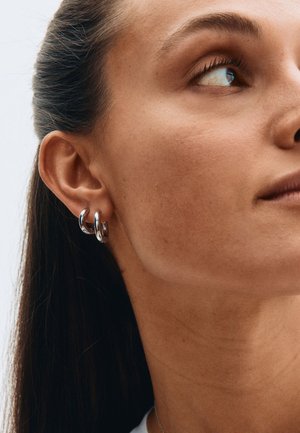 Hõbedased metallist rõngas-kõrvarõngad kahepaikse disainiga, läikiva viimistlusega ja siledapinnalised, kantud mudeli kõrvakilbil.