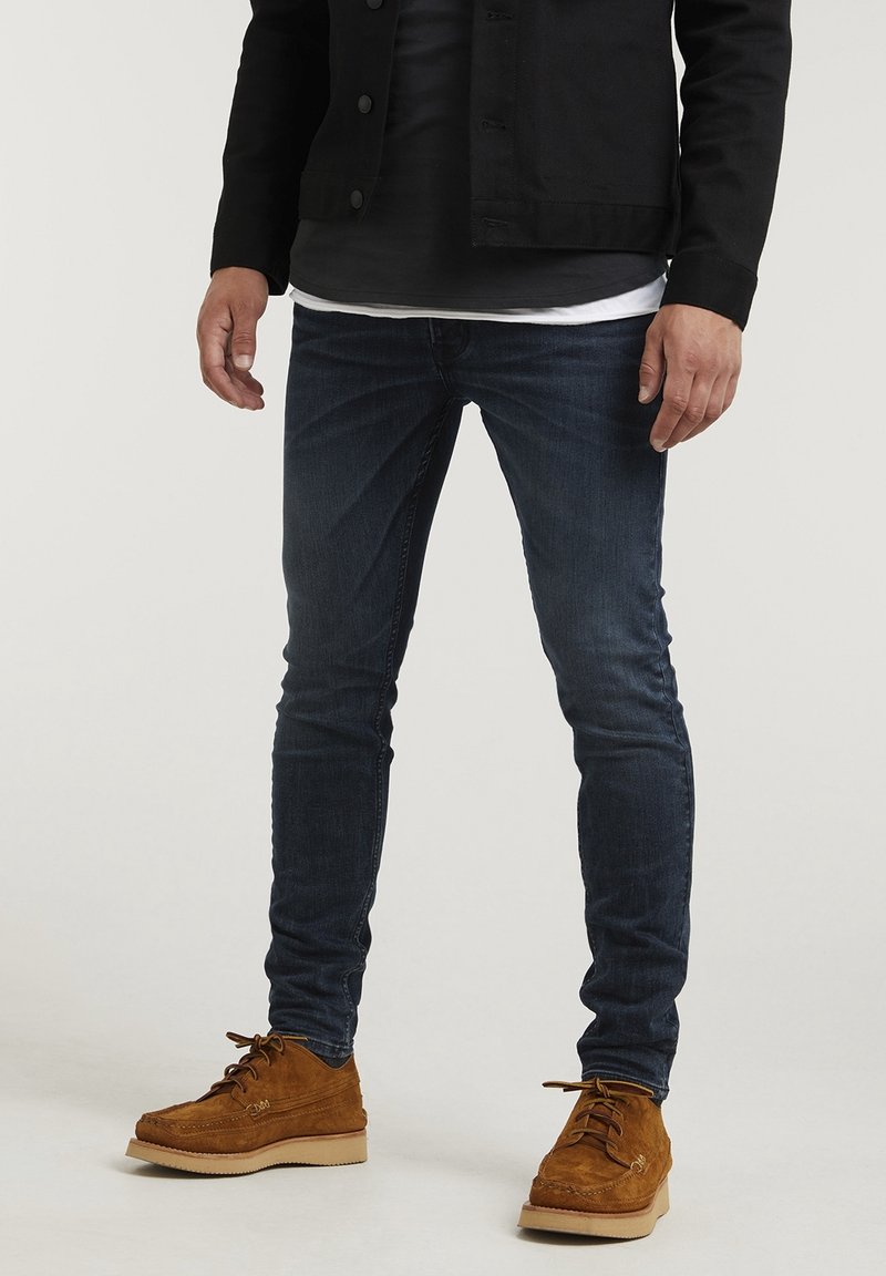 Muž v úzkých tmavě modrých džínách, hnědých semišových šněrovacích botách, černé bundě a s tílky s vrstvami černé a bílé, stojící na jednoduchém pozadí.