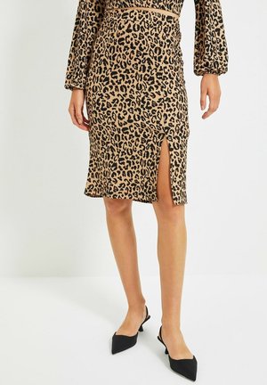 Kvinde iført nederdel med leopardprint med frontslids og sorte spidse slingback hæle, stående mod en ensfarvet baggrund.