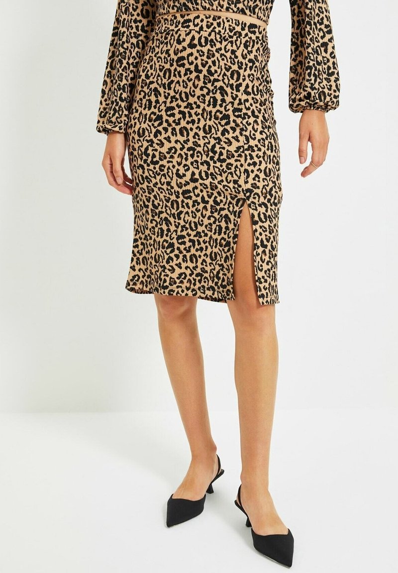 Femme portant une jupe imprimé léopard avec une fente à l'avant et des talons slingback noirs à bout pointu, debout devant un fond uni.