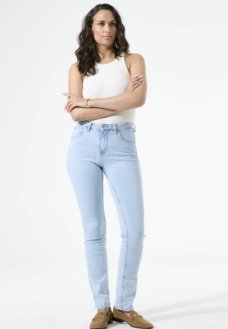 Femme aux longs cheveux noirs portant un haut blanc sans manches, un jean bleu clair et des mocassins marron, debout les bras croisés sur fond blanc.