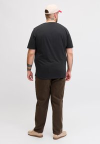 Άνδρας με μπεζ καπέλο, μαύρο μπλουζάκι, καφέ παντελόνι και μπεζ παπούτσια, στέκεται γυρισμένος με την πλάτη προς το φόντο.