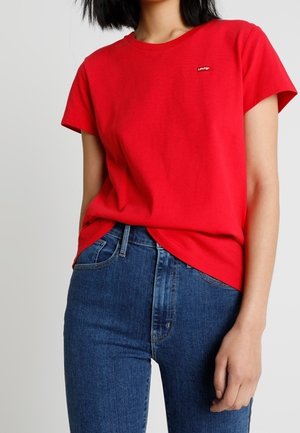 Vrouw draagt een effen rood Levi's T-shirt met korte mouwen, lichtjes ingestopt in een hooggetailleerde blauwe spijkerbroek.