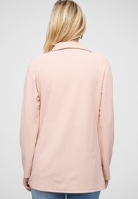Lys rosa langærmet top med krave, blød tekstur og lige pasform, båret med lyseblå jeans, set fra ryggen.