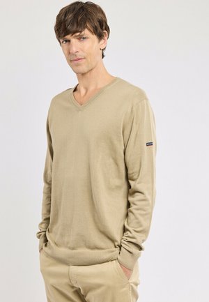 Homme aux cheveux bruns portant un pull beige à col en V et un pantalon kaki, debout les mains dans les poches devant un fond clair uni.