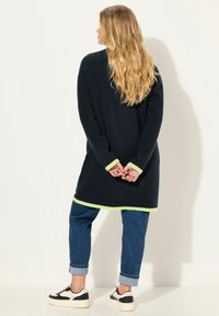 Maglione lungo navy con accenti verde chiaro su polsini e orlo, abbinato a jeans blu arrotolati e sneaker bianco e nero.