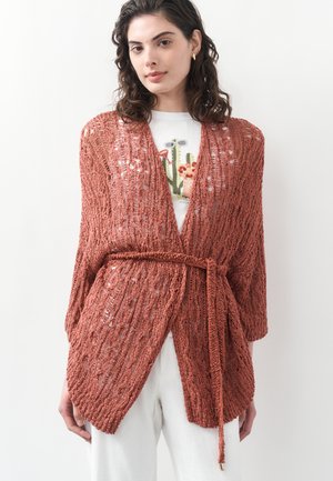 Woman wearing a rust-colored open-knit cardigan with a belt over a white floral-print shirt and white pants, standing against a plain background.