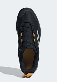 Zapato deportivo negro con tejido de malla, puntera redonda, acentos amarillos y tres rayas blancas. Diseño texturizado y un collar acolchado.