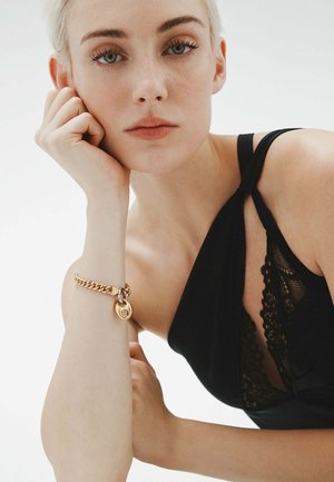 Prenda negra con acentos de encaje, que presenta un diseño asimétrico. Pulsera de cadena dorada con un charm circular en la muñeca. Textura de piel suave.