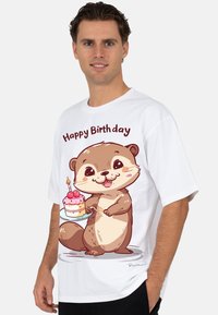 Wit T-shirt met een cartoonotter die een verjaardagstaart met roze glazuur vasthoudt. Tekst erboven: "Gelukkige Verjaardag." Korte mouwen, losse pasvorm.