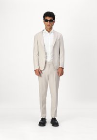 Hellbeige Anzug mit tailliertem Sakko und Hose, kombiniert mit einem weißen Hemd. Schwarze rechteckige Sonnenbrille und schwarze Schuhe runden den Look ab.