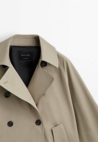 Beige trenssitakki mustilla napeilla, leveillä kauluskäänteillä ja vinosti sijoitetulla etutaskulla, esittää yläosan etu- ja kaula-alueen.