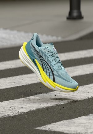 Hellblauer Laufschuh mit strukturierter Oberfläche, schwarzen Streifen, einer leuchtend gelben Sohle und dezenten Markendetails. Schwebend über einem Zebrastreifen.