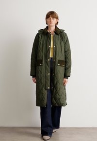 Gequilted olijfgroene lange jas met een capuchon, voorzien van knoopzakken en bruine accenten, gedragen over een lichtgele top en donkere jeans.