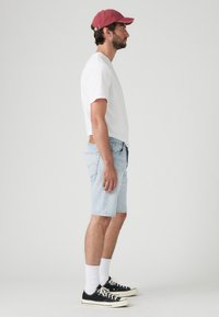 Șorturi din denim bleu deschis, tricou alb și adidași negri cu șosete albe. Căciulă roșie purtată cu spatele. Ținută lejeră pe profilul lateral al unui model masculin.