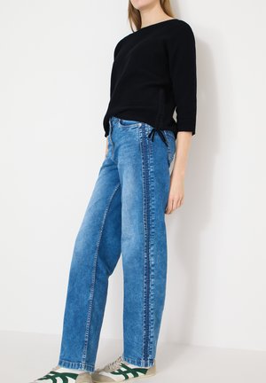 Femme portant un pull en tricot noir, un jean bleu délavé taille haute avec des coutures latérales, et des baskets beige avec des accents verts, debout contre un mur blanc.
