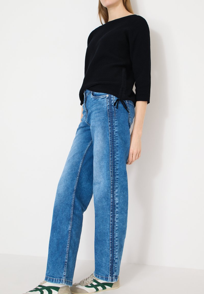Femme portant un pull en tricot noir, un jean bleu délavé taille haute avec des coutures latérales, et des baskets beige avec des accents verts, debout contre un mur blanc.