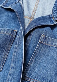 Närbild av en blå denimjacka som visar främre fickor, vikt krage och synliga sömdetaljer.