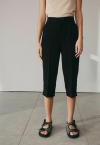 Schwarze, kurz geschnittene Hosen mit vorderem Faltendesign, taillierter Passform, Knopfverschluss und weitem Saum. Kombiniert mit schwarzen, gemusterten Sandalen.