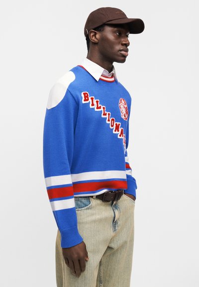 Μπλε πλεκτό πουλόβερ με κόκκινες και λευκές ρίγες, με λογότυπο και γραφικό στην πρόσοψη. Ελαφρύ τζιν παντελόνι και καφέ καπέλο ολοκληρώνουν το σύνολο.