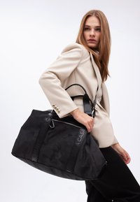 En modell i en beige kavaj håller en stor svart väska med logoband, placerad mot en enkel vit bakgrund. Allvarligt uttryck, stilfull pose.