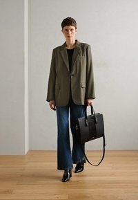 Blazer olive, top noir, jeans bleus larges et sac à main noir avec détail tissé. Chaussures noires à bout pointu, fond de sol en bois.