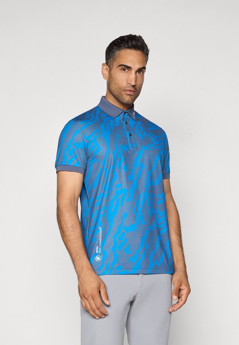 Homme portant un polo à motifs bleus avec le logo "BMW PGA Championship" et un pantalon gris clair, se tenant devant un fond uni.