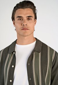 Groen gestreept overhemd met kraag, met verticale crèmekleurige strepen en een gestructureerde stof. Draagt over een wit t-shirt.